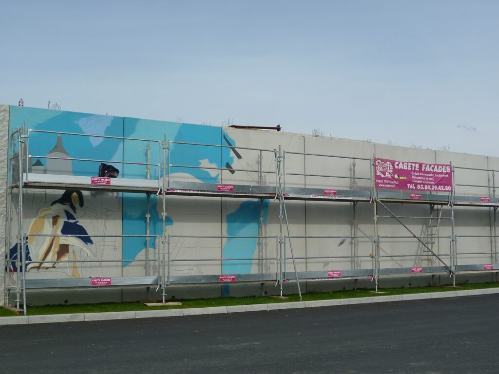 fresque murale en cours de réalisation avec un échafaudage devant. Le mur est en béton gris et il y a de la peinture bleue