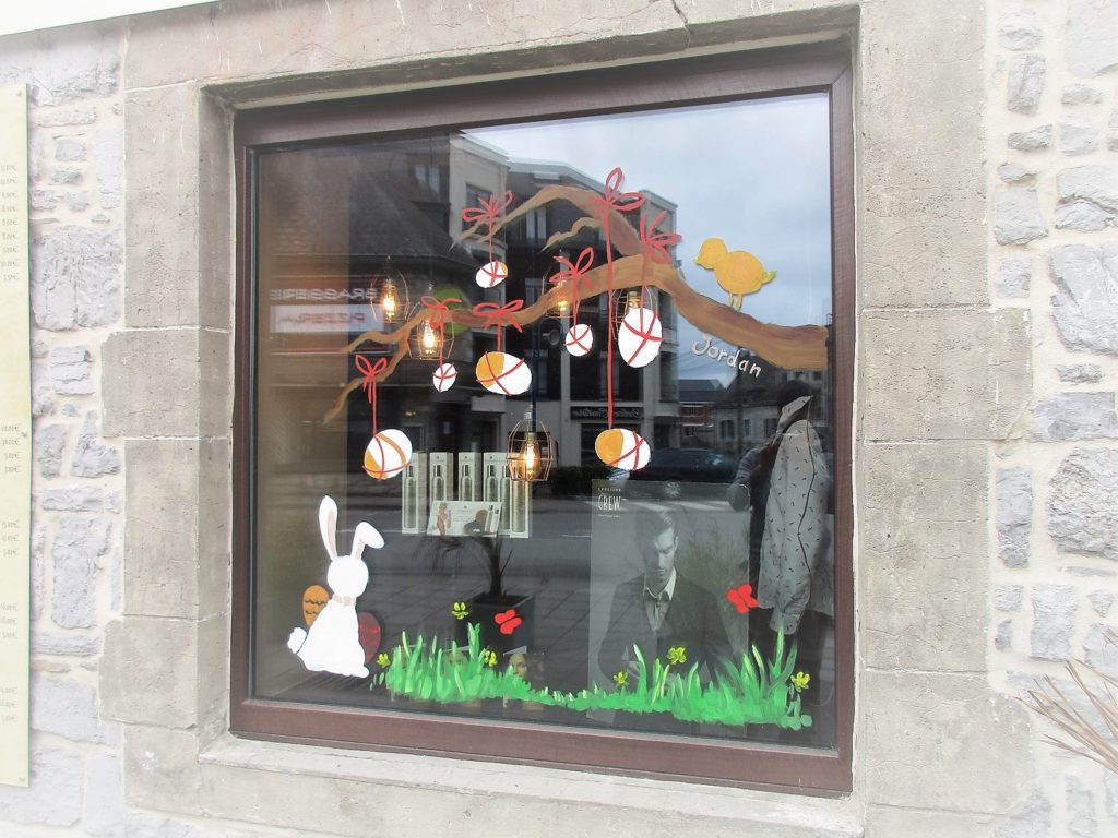 vitrine peinte représentant un lapin et une branche à laquelle sont accrochés des oeufs de pâques. En bas il y a de l'herbe et des fleurs jaunes