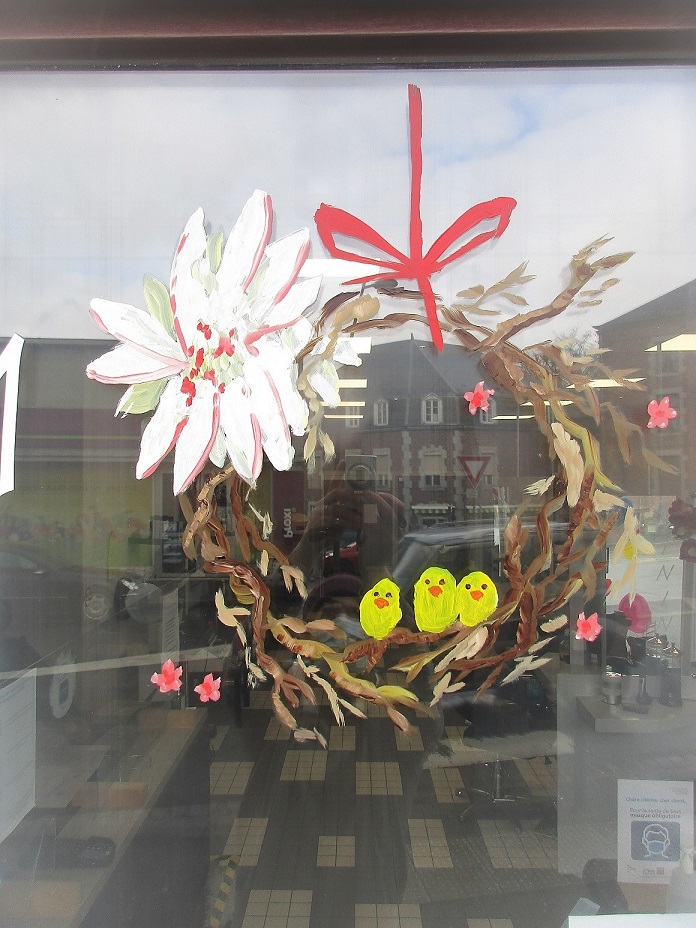 Peinture sur vitrine d'une couronne en bois avec une grosse fleur blanche et 3 poussins jaunes assis. Il y a aussi des petites fleurs roses