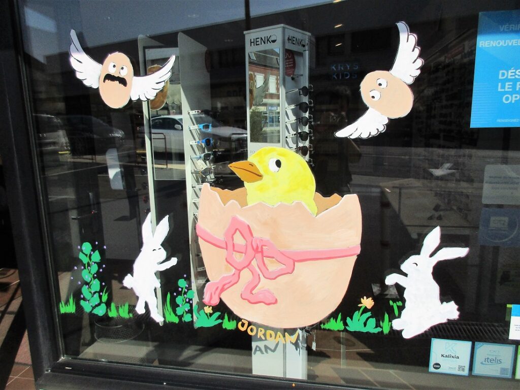 un poussin jaune sort d'un œuf géant, il y a des lapins blancs et des petites fleurs colorées autour, c'est une vitrine peinte de Pâques