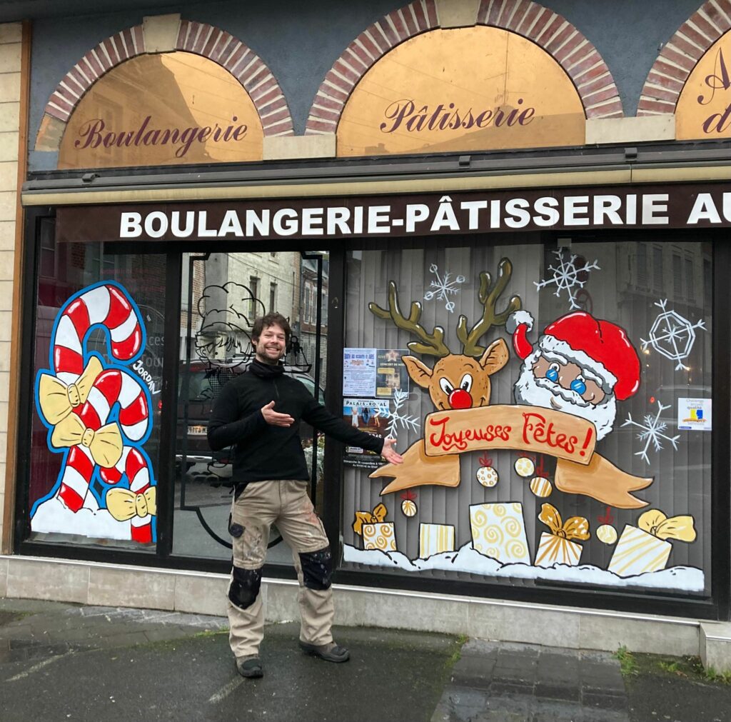 Peinture sur vitrine d'une boulangerie avec Père Noel et rennes. Sura la gauche il y a des sucres d'orge géants et l'artiste pose devant son œuvre