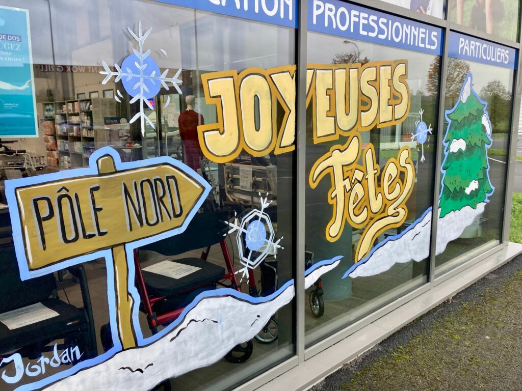 une peinture colorée sur une vitrine avec un panneau indiquant le pole nord, un joyeuses fêtes écrit en jaune et un sapin enneigé. Il y a aussi des flocons en bleu