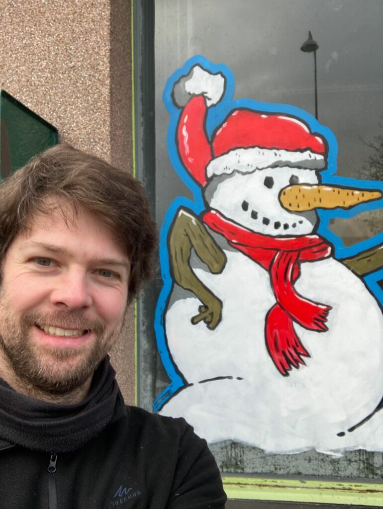 un artiste avec un bonhomme de neige qui a un bonnet rouge et une écharpe dessiné sur une vitre