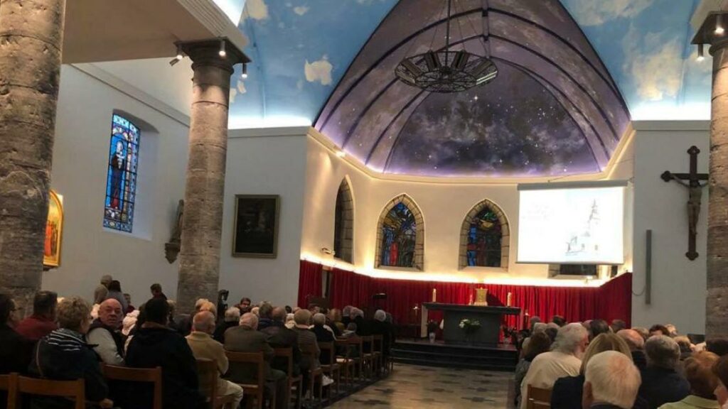 intérieur d'une église remplie de gens assis. Le plafond est peint en ciel étoilé et nuageux. Il y a des grosses colonnes en pierre